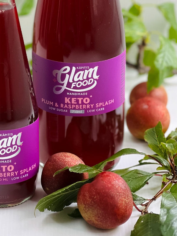 Two bottles of Glam Food plum and raspberry splash on a white surface with plums and green leaves.